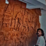 Large, terracotta-colored fiberglass bas-relief mural depicting a historical procession from Hampi. An artist, looking toward the viewer, stands in the foreground to the right, emphasizing the mural's immense scale.